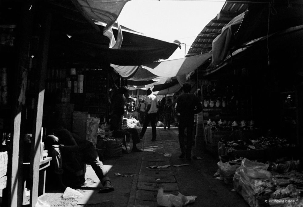  A black and white photograph taken at hip level, showing a bustling market alleyway. People walk through the narrow passage lined with stalls covered by awnings. Various goods appear to be for sale on either side, though the details are somewhat obscured by the contrast and shadows. The ground is uneven and scattered with debris. Light filters in from the open sky above the awnings, creating areas of bright contrast with the deep shadows.