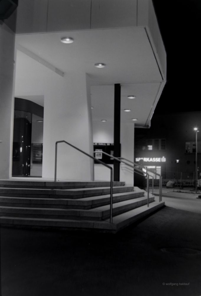 A black and white photograph captures the illuminated entrance of a building at night. The architectural lines of the steps and overhead canopy are emphasized by the contrast of light and shadow, with a subtle glow from the "SPARKASSE" sign in the distance.