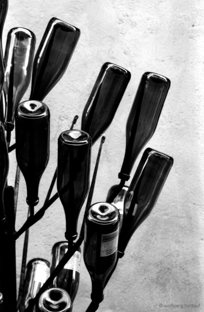 This black and white photograph captures a close-up view of a metal wine bottle rack filled with several empty wine bottles. The rack is positioned diagonally across the frame, creating dynamic lines. 