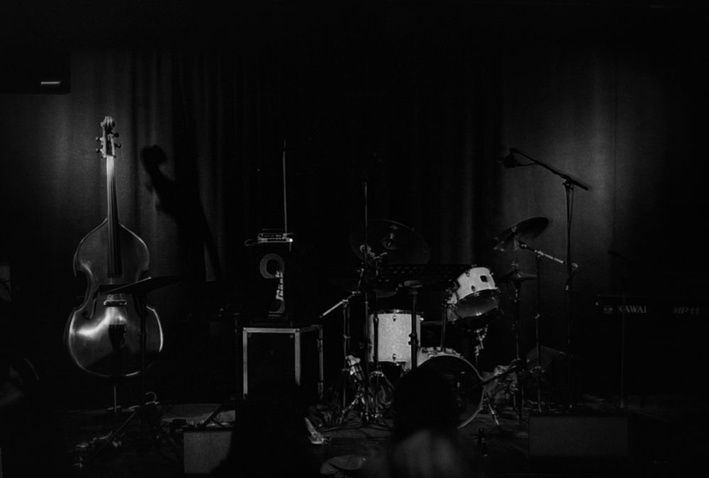 An analog black and white photograph captures a stage moments before a jazz concert. The dimly lit scene features an upright bass standing prominently on the left, its reflective surface catching the faint light. To its right, a drum kit and a microphone stand are set up, awaiting the musicians. The overall atmosphere is one of quiet anticipation, characteristic of the calm before a live performance.