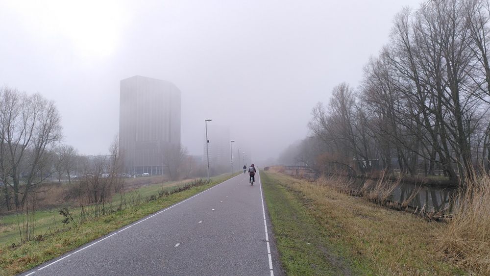 A dyke with cyclists and fog in the distance. A tower is partially covered by the fog.