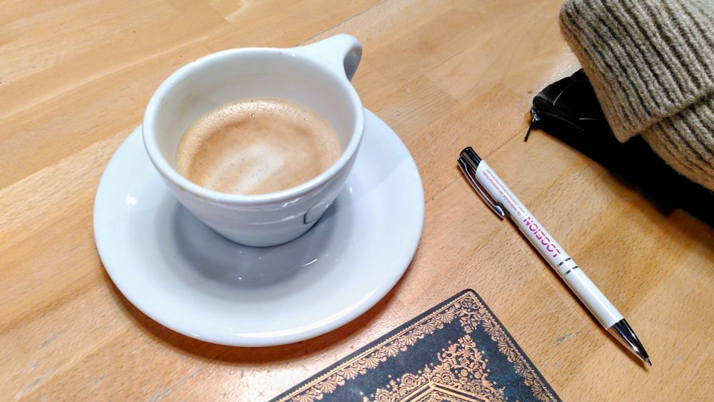 White cup and saucer with a latte on a table, parts of a notebook, pen and hat are visible. 