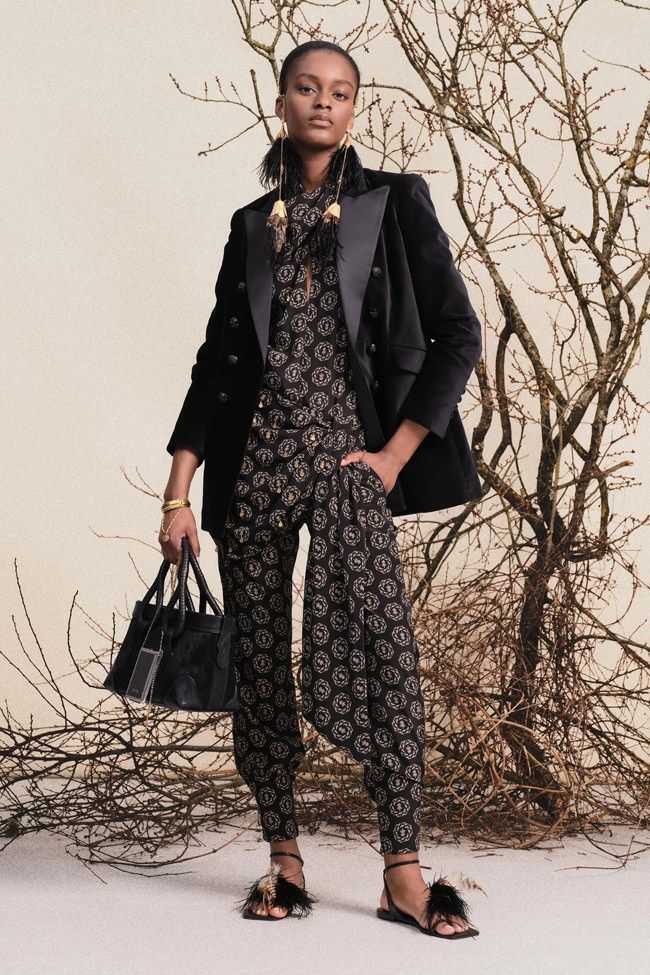 Model wearing a black jumpsuit with black velvet coat, black purse, and black sandals.