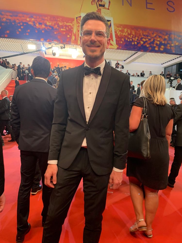 Snapping that disallowed red carpet selfie at the 2019 edition of Cannes film festival. I’m wearing black tie suit with bow tie in front of the red steps. It’s night time and all the lights are on so the shadows go every direction and things seem to sparkle. 