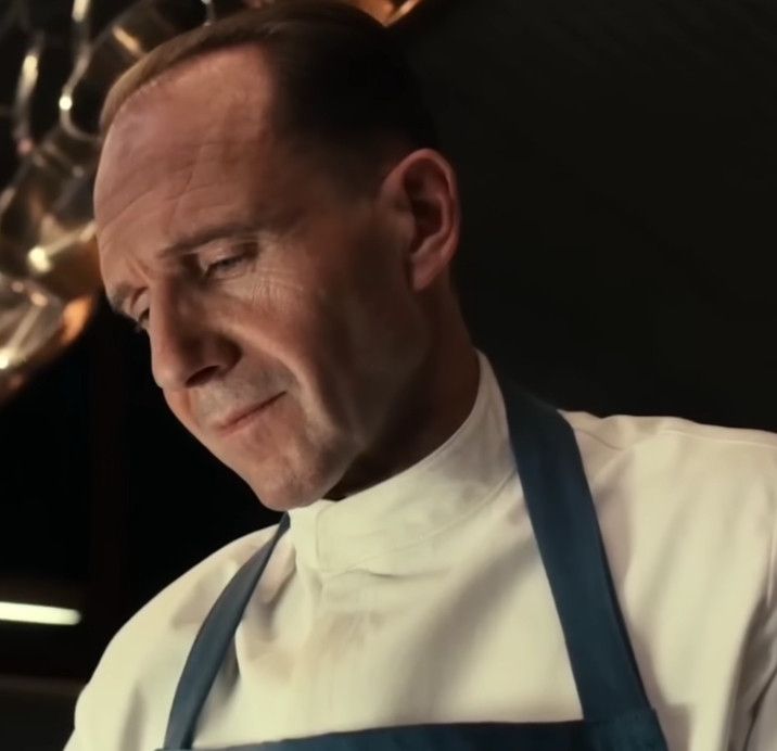 The main chef from the movie the Menu smiling downwards while making a cheeseburger 