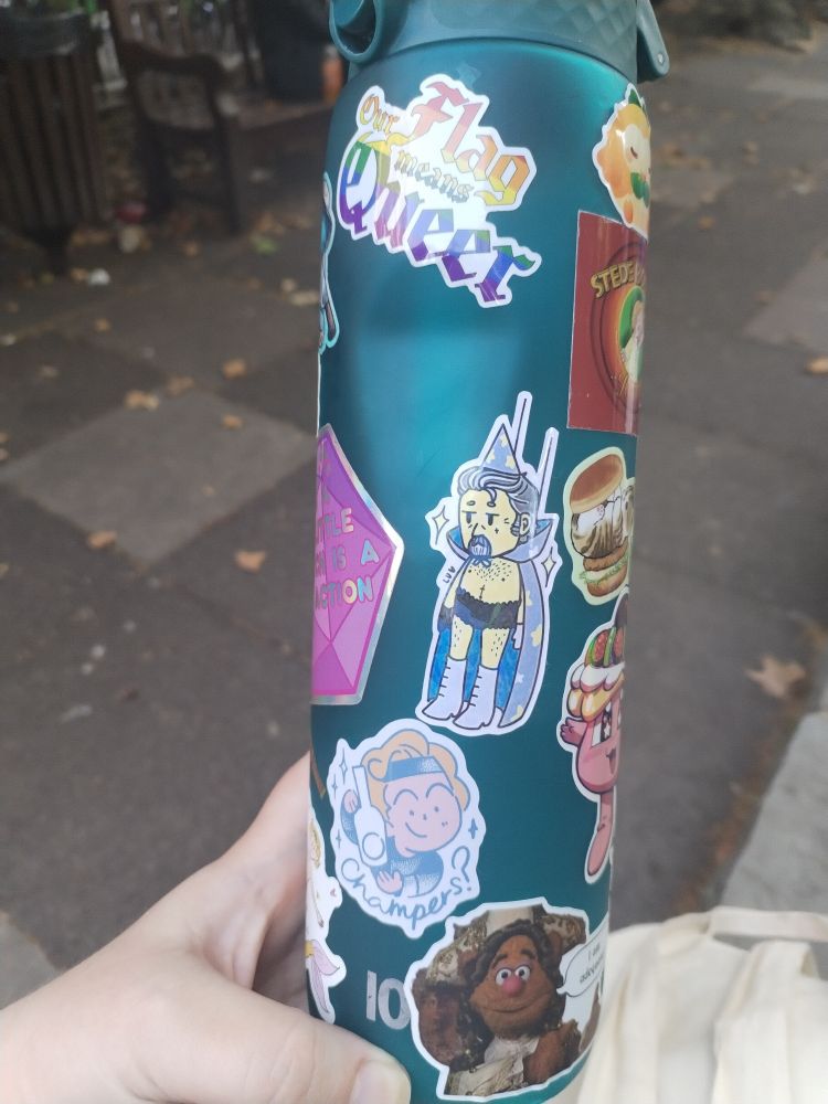 A teal water bottle covered in stickers, including the OFMD logo but it says Our Flag Means Queer in rainbow flag colours, Izzy hanging from stunt wires like the SpongeBob meme, Stede asking "Champers?", and Fozzy Bear in Muppets Treasure Island saying, "I am adequate!"