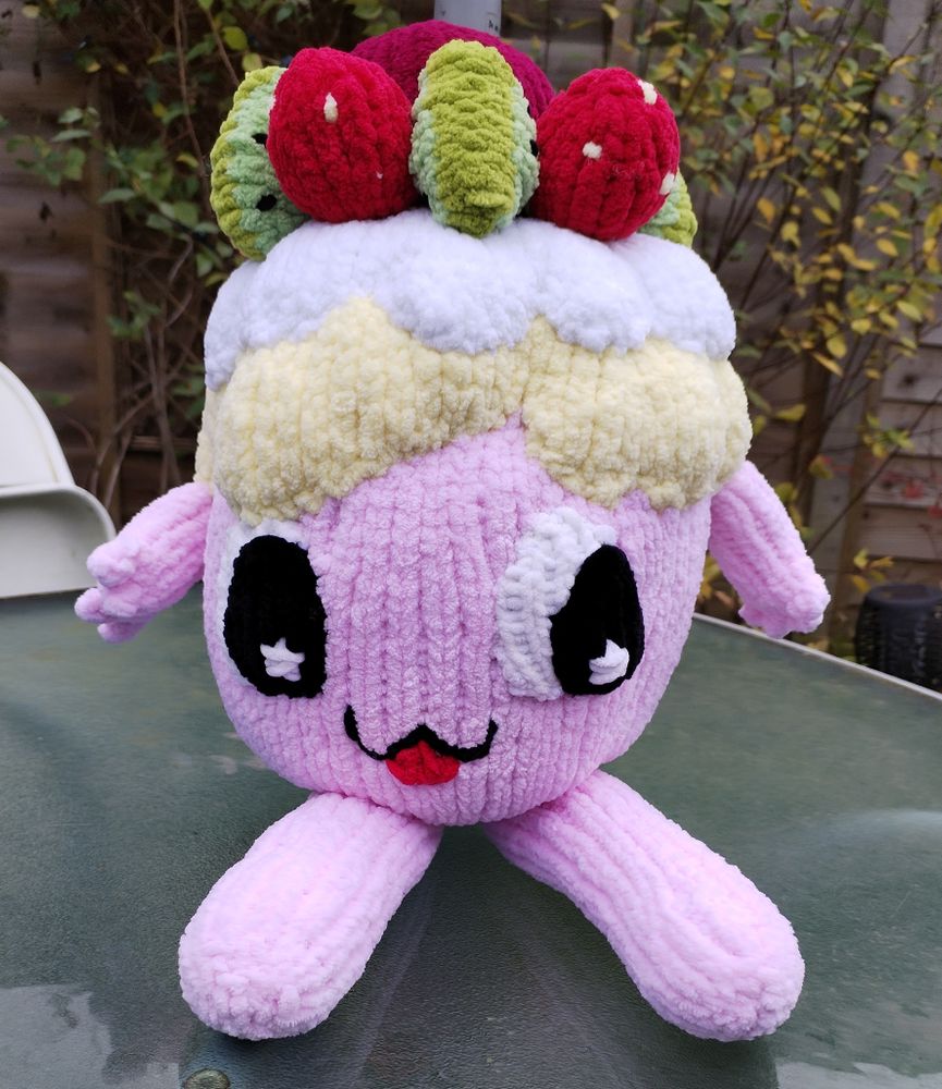 A knitted plushie of Pabu-Chan - a pink character with long legs, little arms, and on his head there is a layer of meringue, a layer of cream, and on top a ring of kiwis and strawberries and a big cherry in the middle. She's sitting on a glass table outside, legs sticking out. 