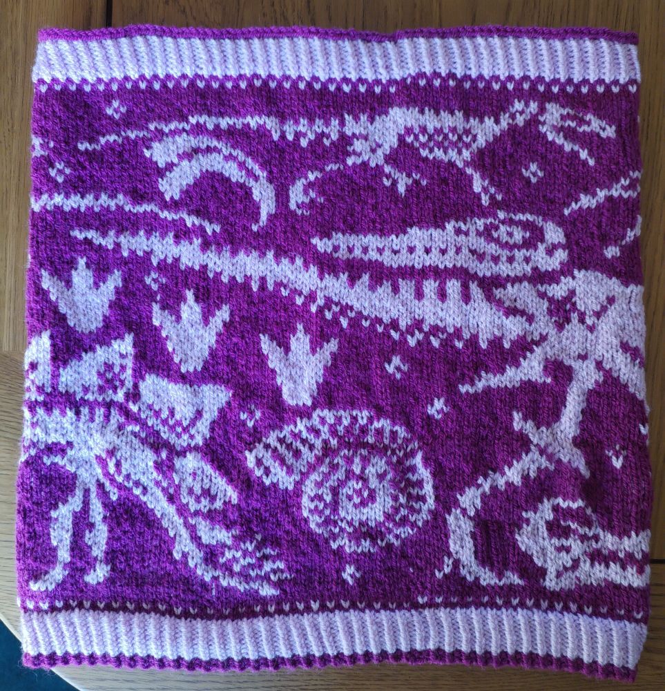 A photo of a knitted cowl in dark and light pint with dinosaur fossils across it. This photo features an ammonite, a raptor claw, footprints, back halves of a stegosaurus and t-rex, pterodactyl skull and a tiny raptor-esc dinosaur I've forgotten the name of 