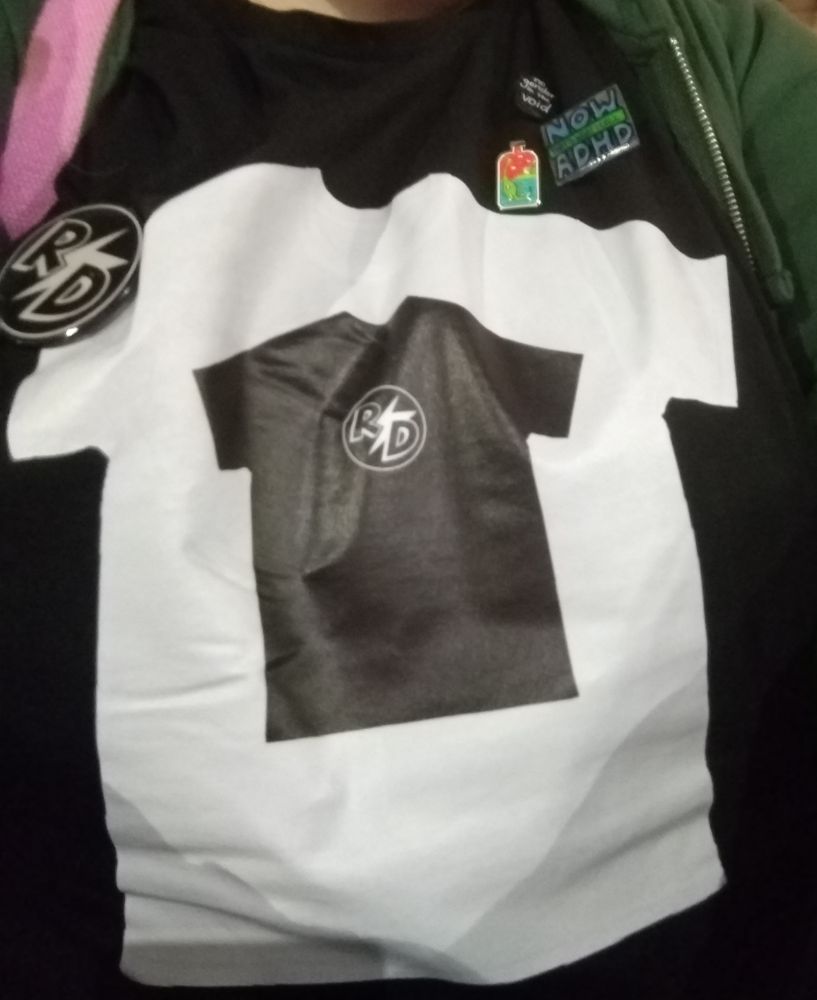 A photo of me wearing a black t-shirt with a white t-shirt printed on, which has a smaller black t-shirt inside that with the Rhys Darby logo. I'm also wearing a R⚡D badge, a nessie in a bottle pin, a pin saying Now That's What I Call ADHD, and a little black pin saying "no gender in the void".