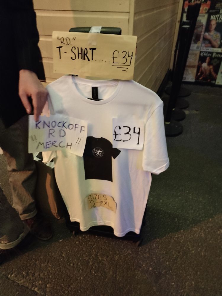A photo of a white t-shirt with a print of the Rhys Darby logo t-shirt. Handmade labels are stuck on it saying "RD" t-shirt, £34, knock off RD "merch", sizes S-XL