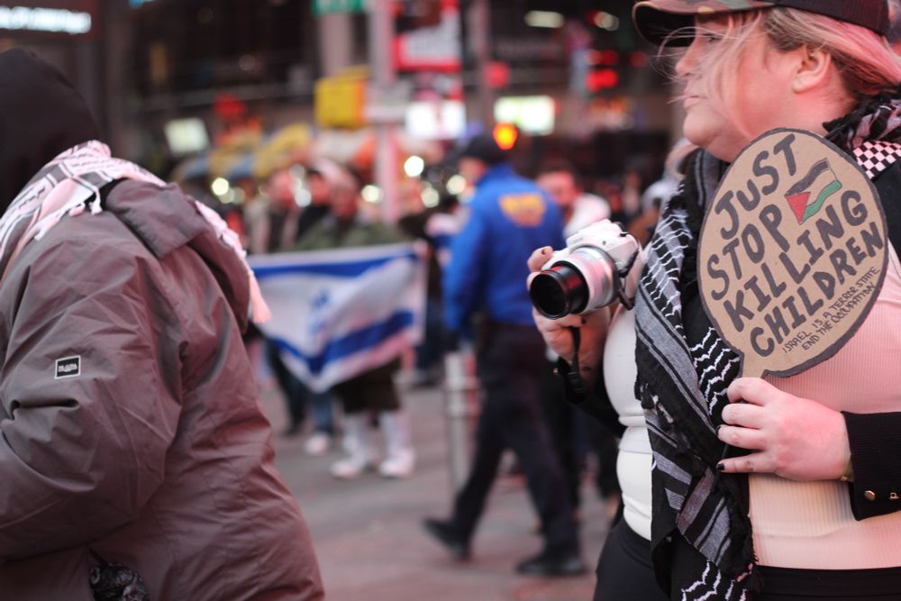 People hold an Israeli flag in the background and in font of them people walk with a woman wearing a black Keffiyeh holding a camera and a round sign saying “just stop killing children” with a Palestinian flag on it