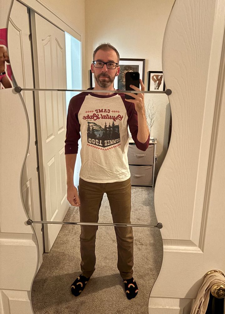 A gay man taking a picture of himself in the full length mirror on the back of his door. He’s very tired and wearing jeans and a white and red semi-long-sleeved shirt with some Camp Crystal Lake graphic and rainbow socks. 