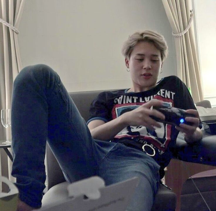 Blond Jimin in a graphic t and jeans, manspreading on a chair as he looks at a camera (?). Who cares what's in his hand, that's not what we're focused on anyway.