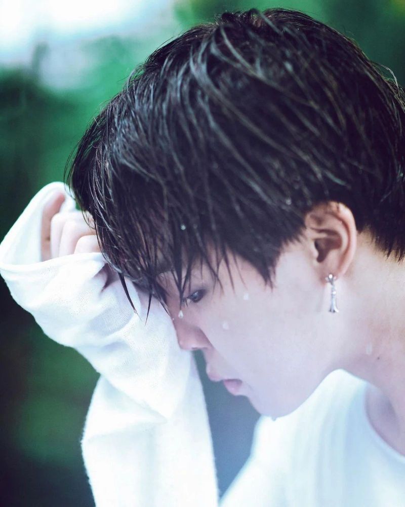 Close up of Jimin's profile. He is wearing a long sleeved white shirt and is looking down, one hand to his forehead. It is raining.