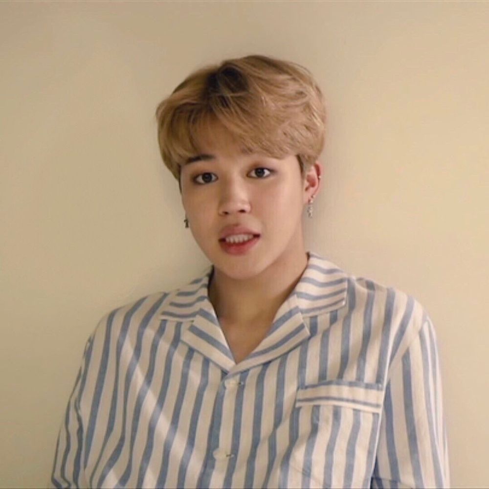 Blond Jimin in a blue and white striped pajama like shirt making a cute face as he stands in front of a blank wall. 