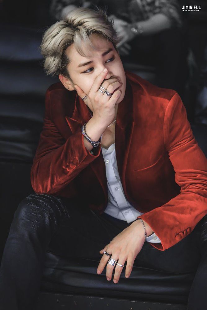 Blond Jimin in tight black pants, a white button up and what looks like a crimson jacket made of velvet. He has one hand up by his face and the other is between his legs...