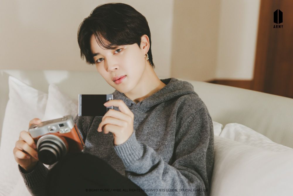 Jimin sitting on a white couch holding a camera and a polaroid. He is wearing a soft looking gray sweater and dark pants.