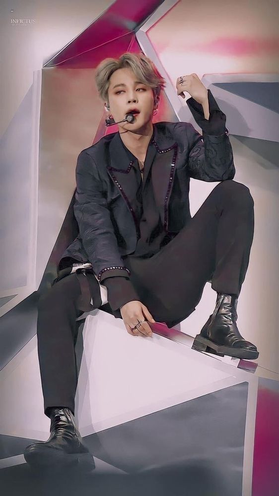 Jimin in a black suit sitting on a geometrical type shape, slightly manspreading.