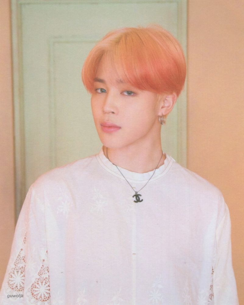 Pink (almost orangey) haired Jimin in a white shirt standing in front of a yellow wall and green door.
