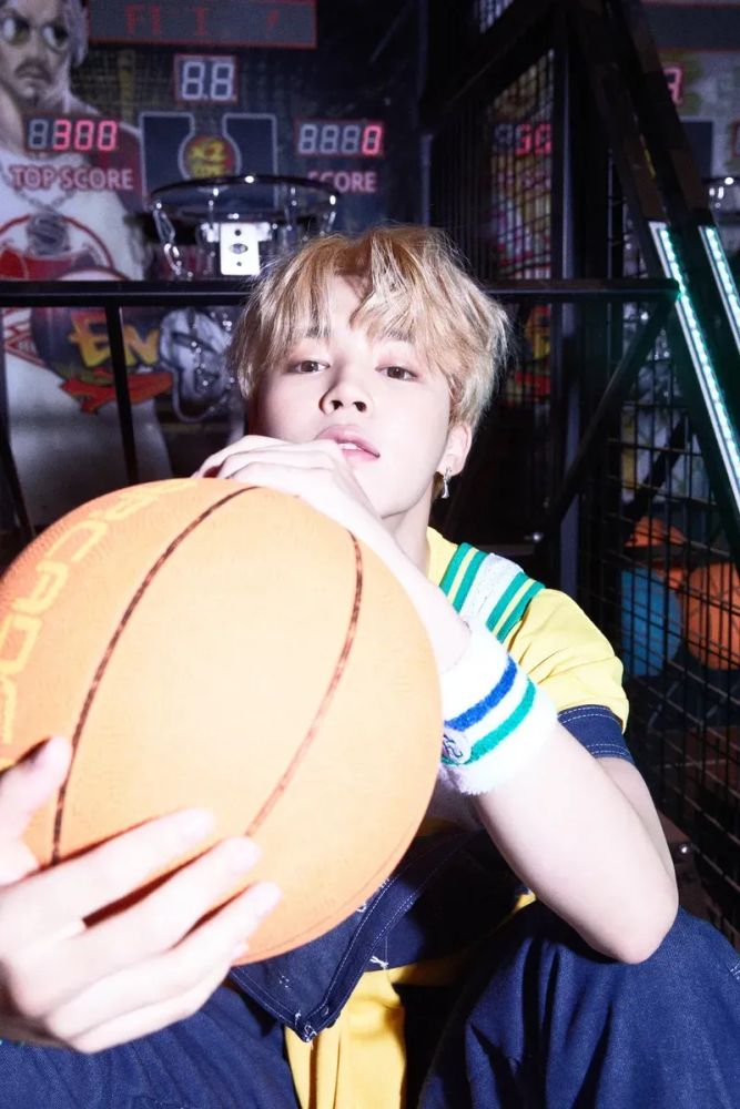 Blond Jimin holding a basketball as he sits in front of a basketball arcade game.