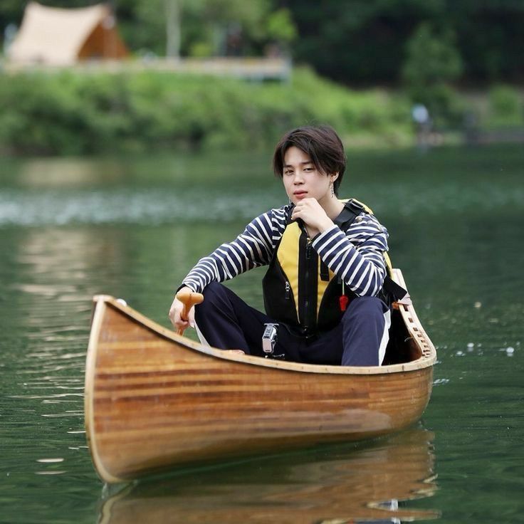 Jimin in a striped long sleeve shirt sitting on a small canoe in the middle of a lake. 