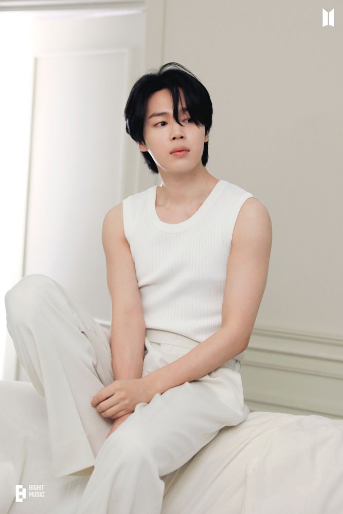 FACE era Jimin with black hair wearing a white tank top and light beige pants sitting on the back of a couch looking to the right.