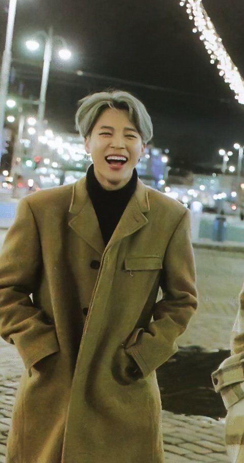 Jimin in a beige trench coat smiling/laughing. 