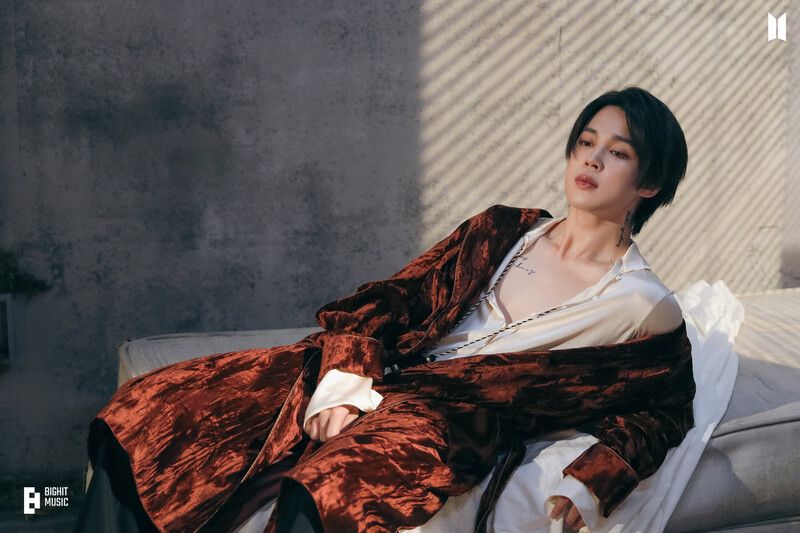 Jimin lounging on a mattress wearing a burgundy robe over a silk shirt that's showing off half his chest. 