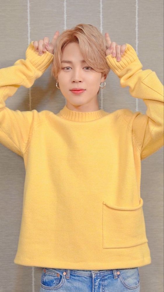 Jimin in a bright yellow sweater, holding his hands up on either side of his head.