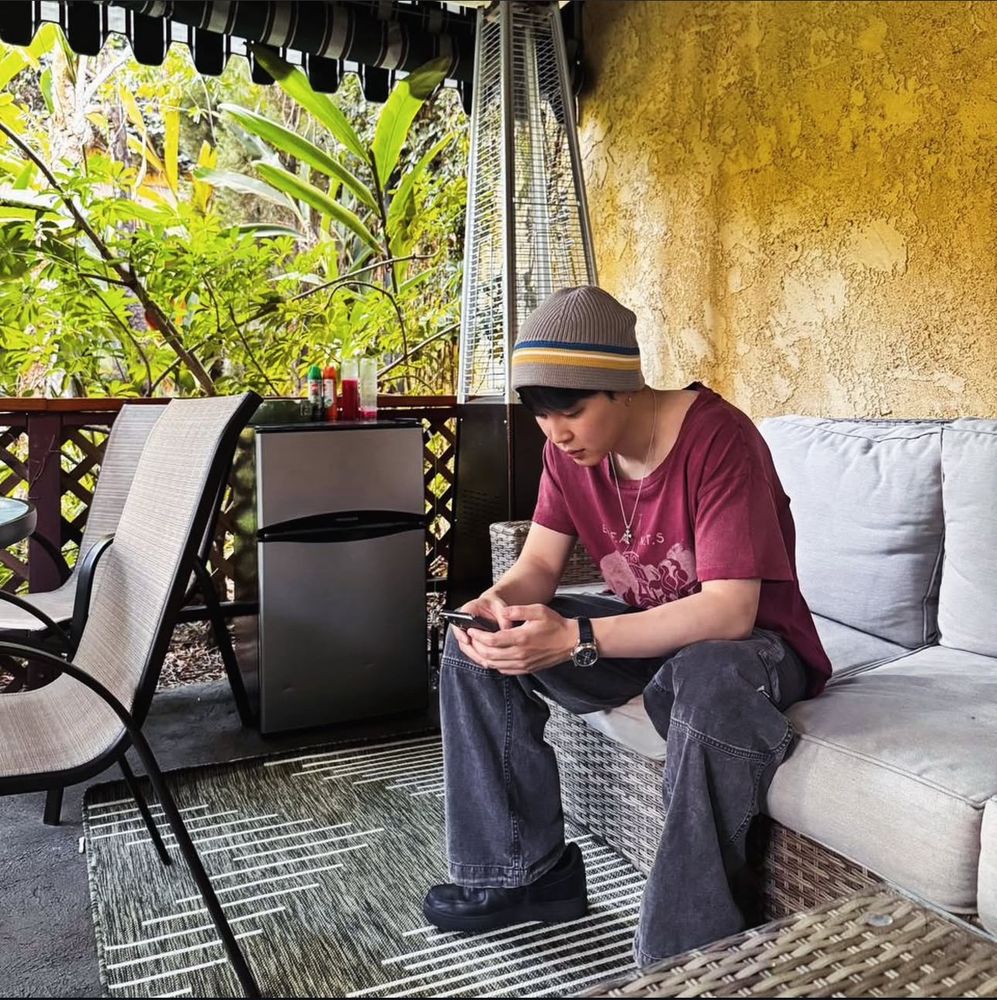 Jimin sitting on a patio set outdoors in jeans, a red tshirt and a gray beanie. He’s looking at something on his phone. 