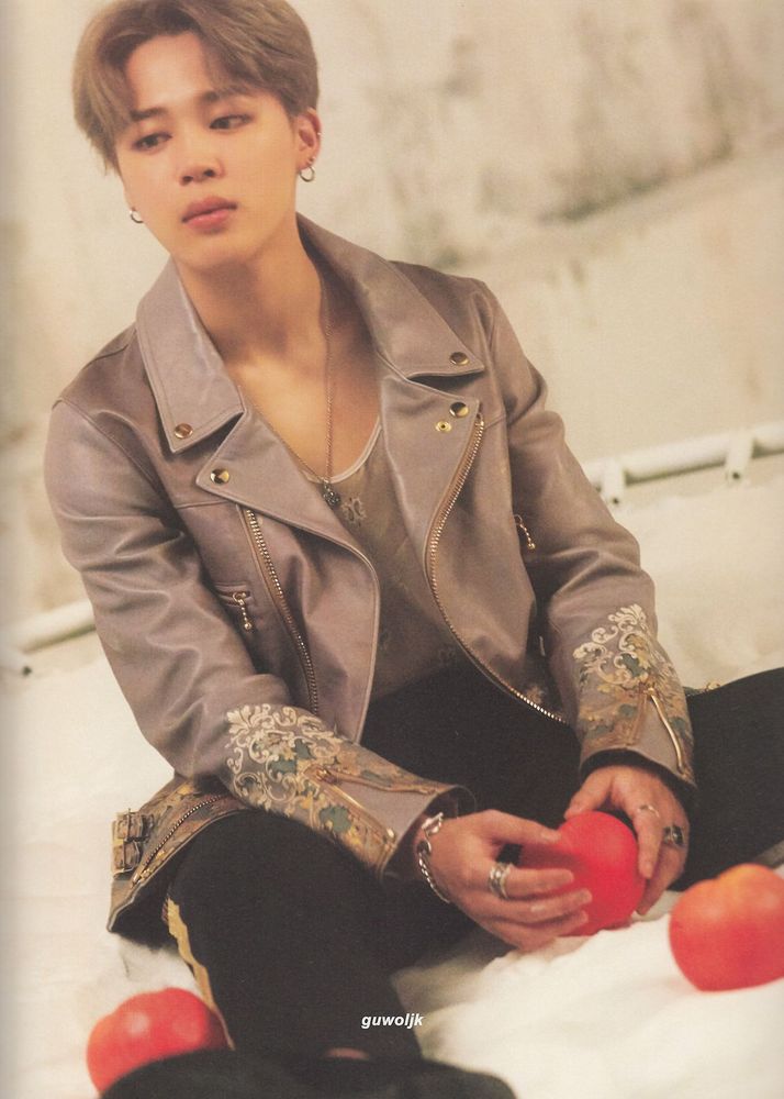 Jimin in black pants and a gray leather jacket sitting on the floor holding an apple in his hand. There are a couple other apples on the floor by his legs, too.