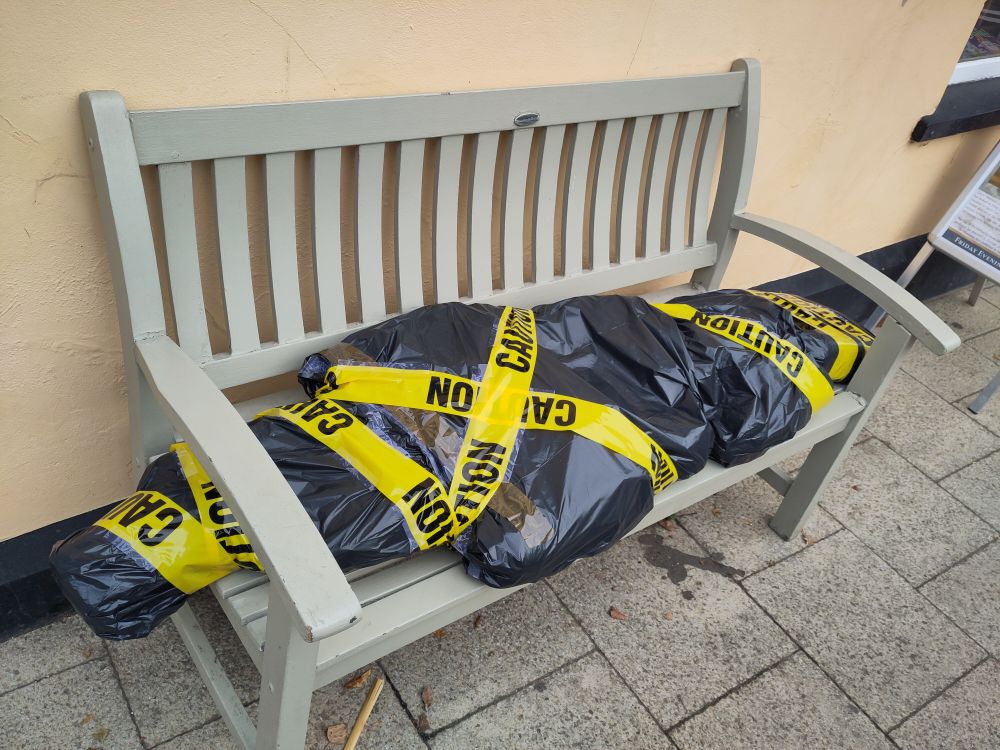 A scarecrow made to look like a body in a taped-up body bag, on a bench 