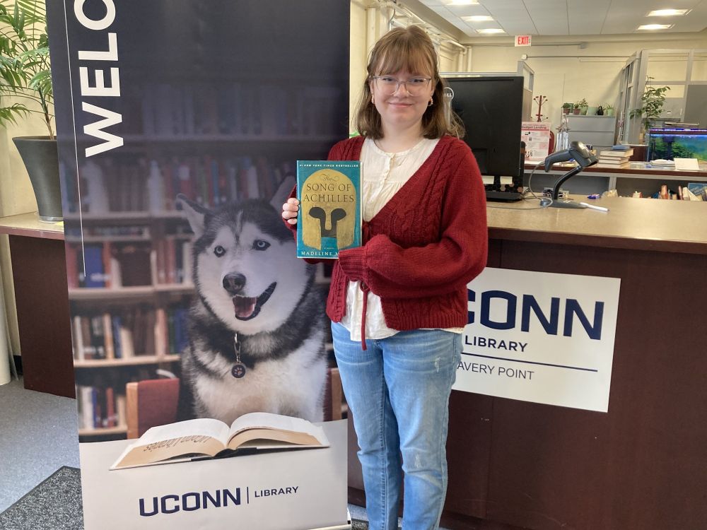 Student Elena Smith holding the book "The Song of Achilles" by Madeline Miller in front of the reference desk next to the Welcome sign with Jonathan the Husky.