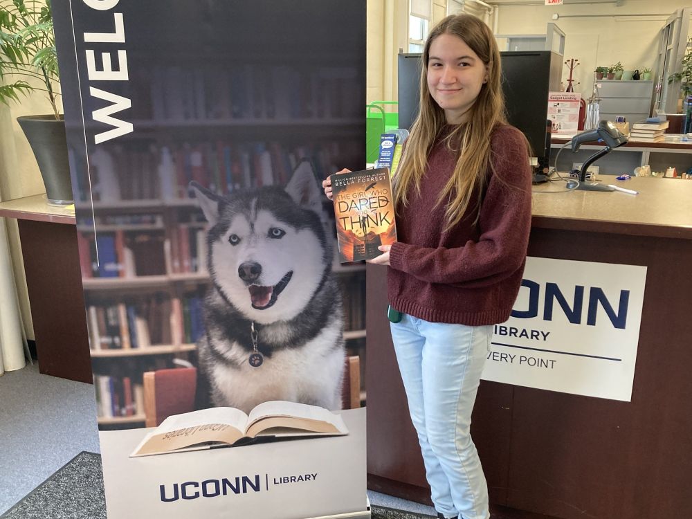 Student Alyssa Spellman holding the book "The Girl Who Dared to Think" by Bella Forrest in front of the reference desk next to the welcome sign with Jonathan the Husky.