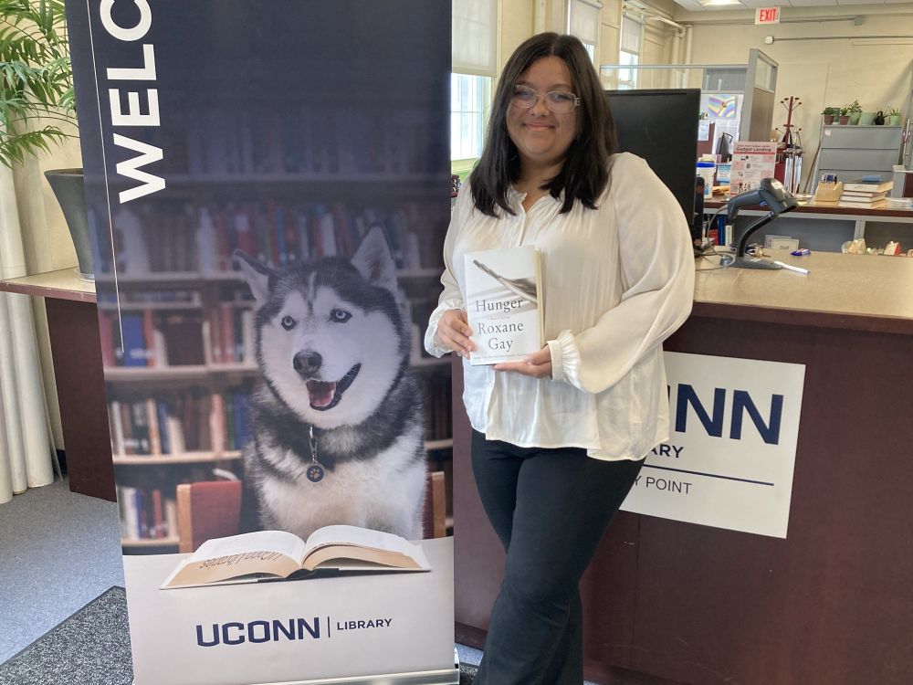 Student Katelyn Lamprey holding the book "Hunger: A Memoir of (My) Body" by Roxane Gay in front of the reference desk next to the welcome sign with Jonathan the Husky.