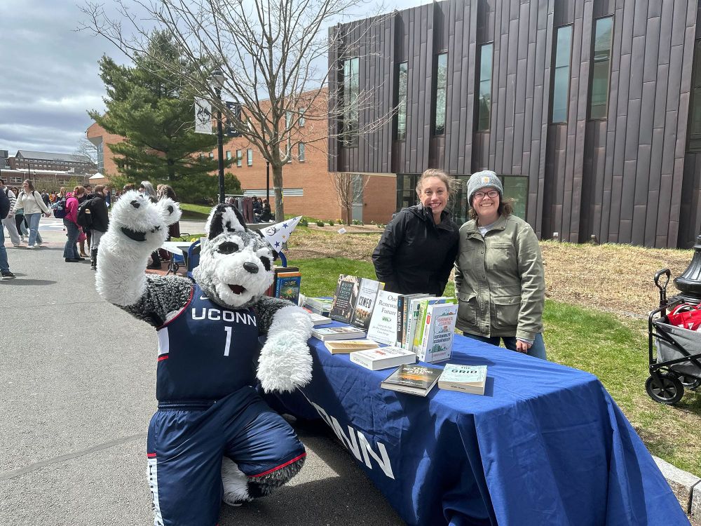 Photo of Jennifer Schaefer, Katie Fiducia and Jonathan outside on Fairfield Way at the UConn Storrs Campus next to a table with books on it.