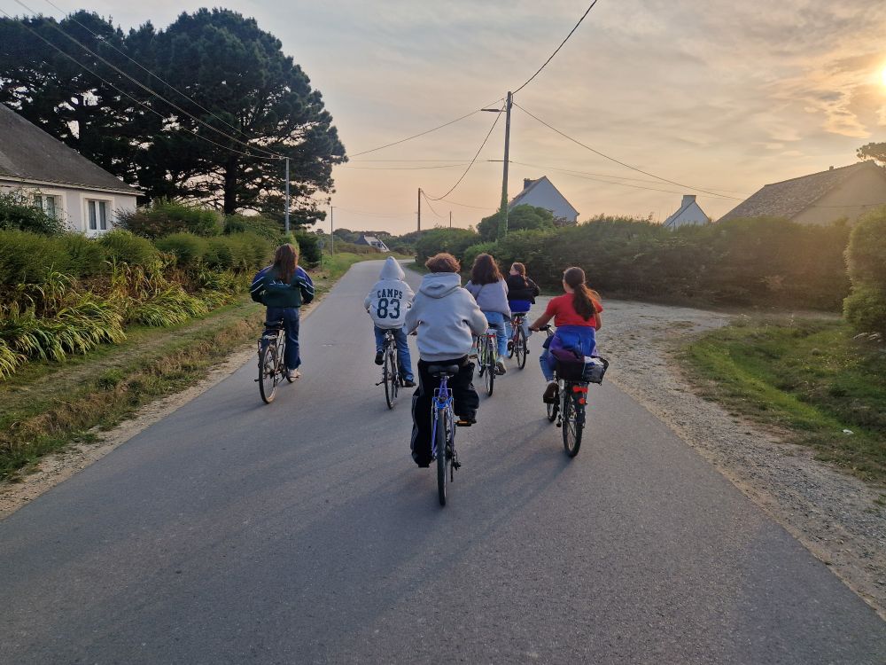 Photo d'un groupe de 6 ados à vélo. Le soleil se couche dans le ciel. Maisons bretonnes sur les côtés 