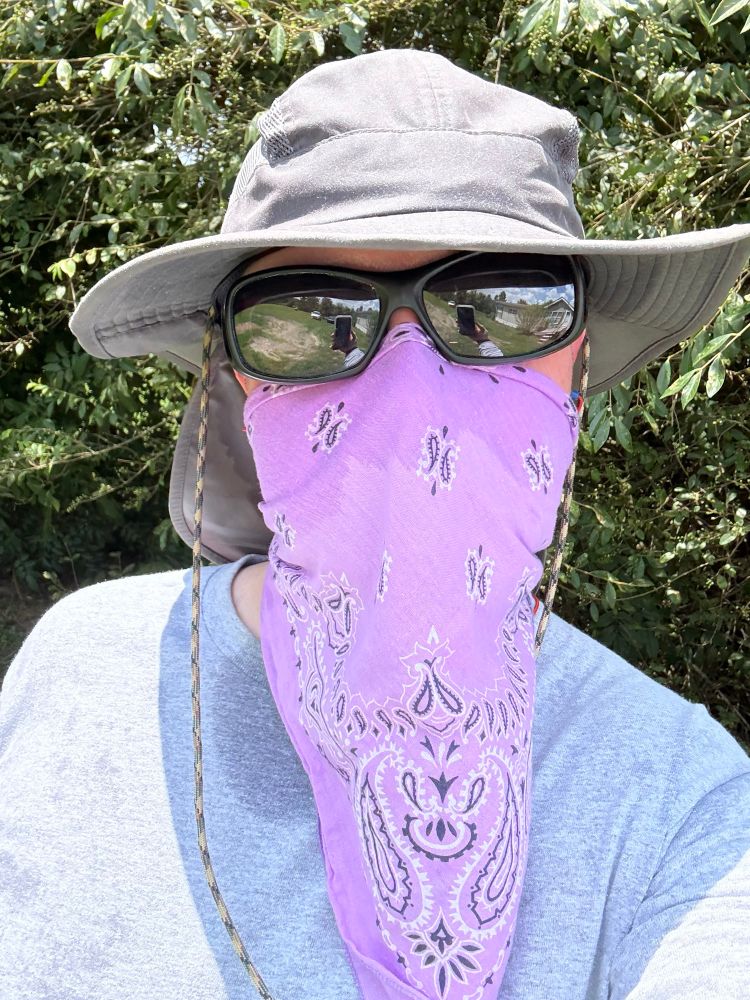 A future with a long lavender colored bandana covering the lower half of their face and neck. They’re wearing a grey longsleeve shirt, large black sunglasses, and a wide grey hat. 
