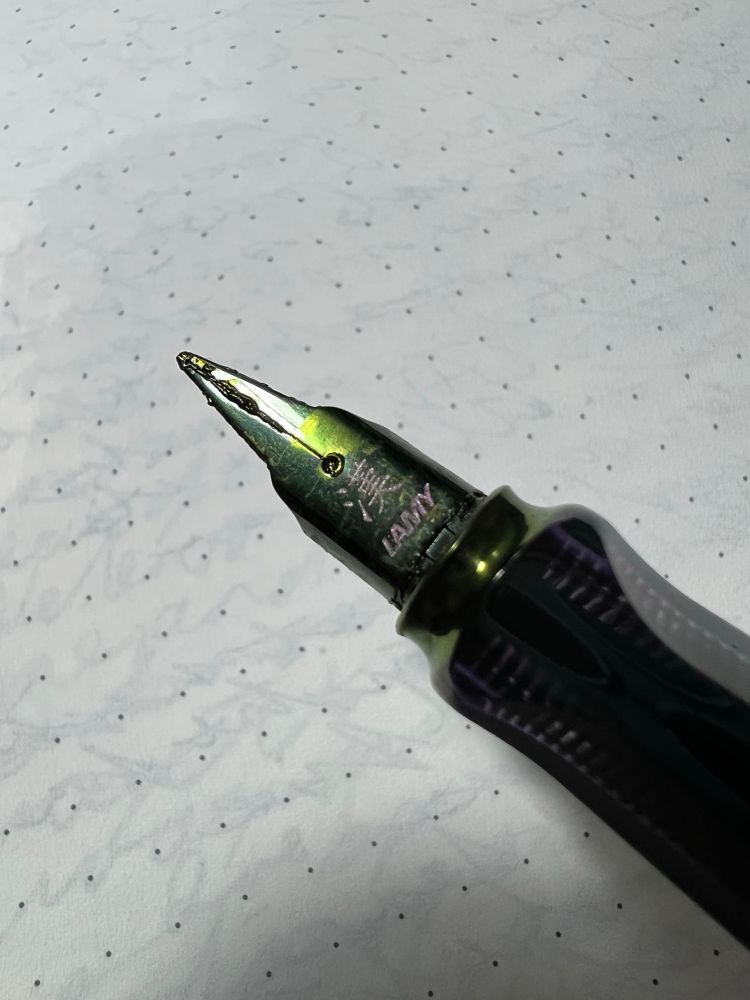 Closeup of a Lamy LX with the cursive nib. The nib itself is black and there is a green sheen across it thanks to the power of Lamy Dark Lilac 2024, which sheens and cruds up at the drop of a hat. 