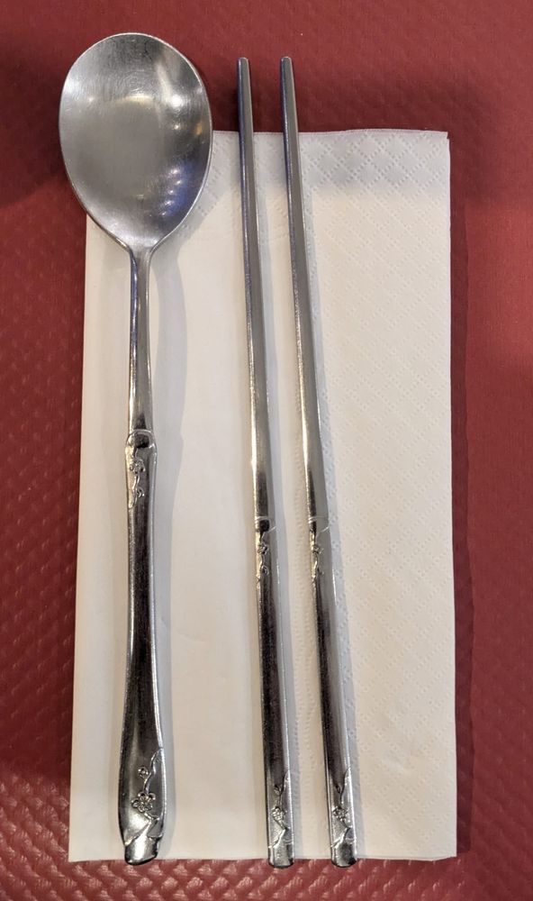 Stäbchen und ein Löffel aus Metall liegen auf einer weissen Serviette bereit.
