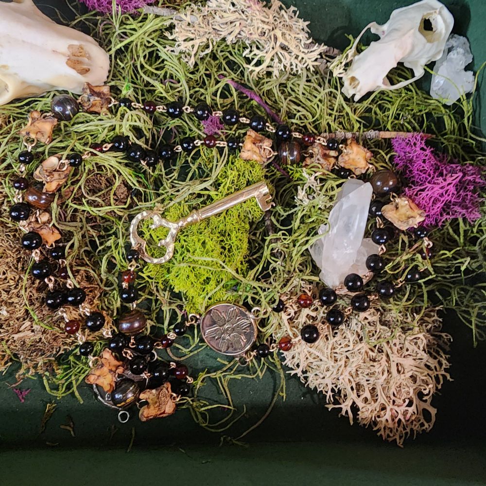 Prayer beads featuring my signature Eye and Six Hands devotional Hekate talisman and wardrobe key cast in bronze, strung on bronze wire, with genuine rattlesnake vertebrae and beads of tiger iron, jet, and amber. The total bead count is: six 8mm tiger iron beads, ten 10mm rattlesnake vertebrae, thirty-two 3mm jet beads, fourteen 2mm jet beads, and fifteen 2mm amber beads, for a total of seventy-seven beads.