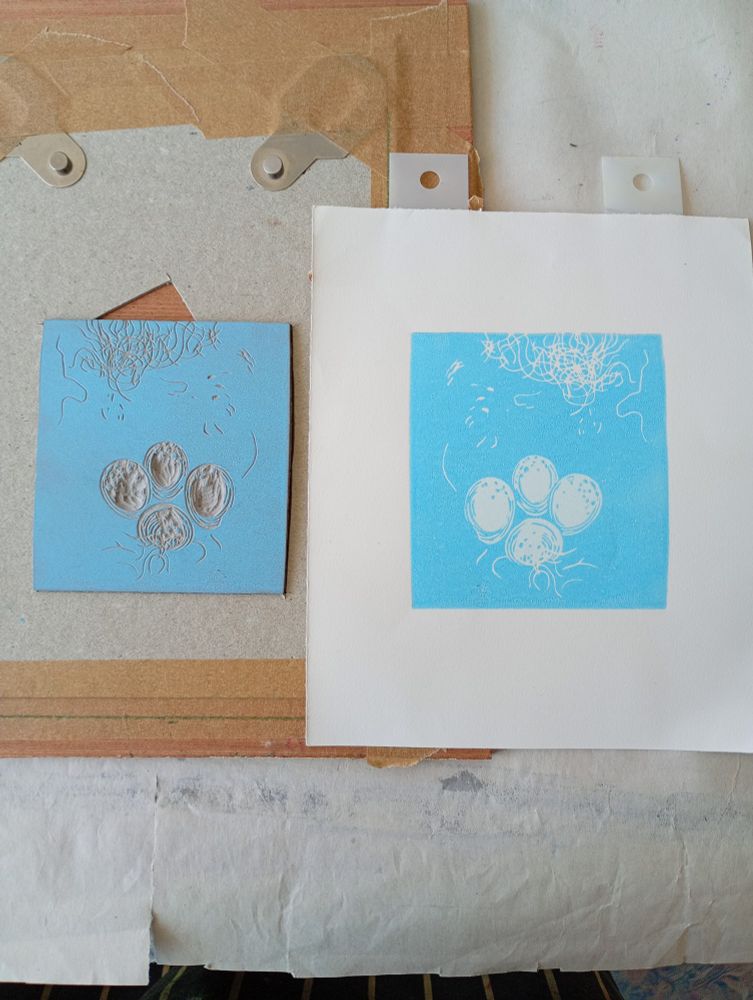 2nd layer of a lino reduction print - a very pale transparent blue covered with the darker eggshell blues of the egg shadows. The photo shows the block in its registration system of a closely cut to fit grey board and Ternes Burton pins next to the printed second layer.