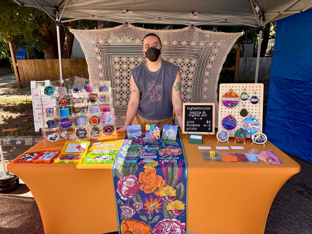 Juniper standing behind the table of her vending booth. She’s mid-size with a buzzed head and arm tattoos, and wears round silver glasses, a black N95, a grey muscle tank and brown shorts. Her table is covered in a golden wrap with a colorful floral runner in the middle. There are stand up displays of stickers and embroideries, a price menu, prints, bins of Pride pins, a spread of cross stitch patches, and business cards. Her backdrop is a large crochet piece.