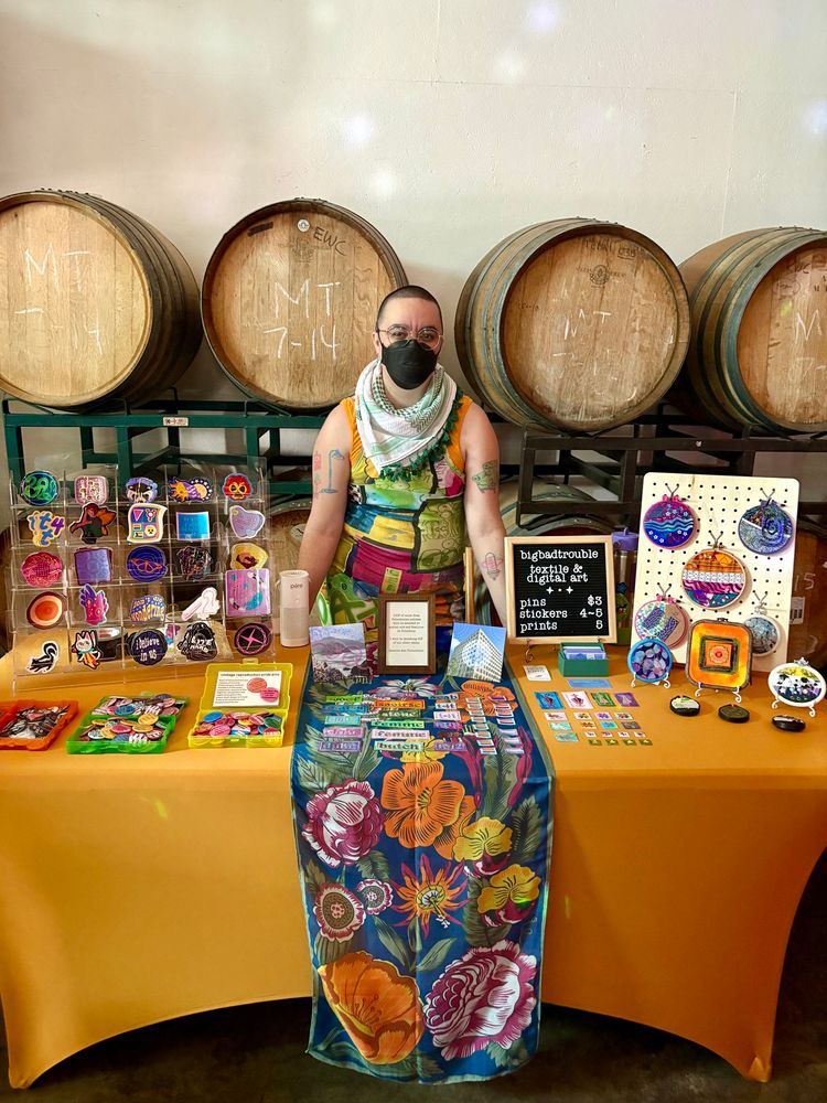 Juniper standing at her vending booth inside a winery. She’s wearing a black N95 mask, a kuffiyeh in the colors of the Irish flag, and a colorful bodycon dress. Her booth is covered in a gold tablecloth with a colorful floral runner. From left to right, there’s a clear acrylic sticker display, bins of repro Pride pins, square prints, cross stitch patches, a price menu, business cards, and displays of embroideries, cross stitches and fabric collages.