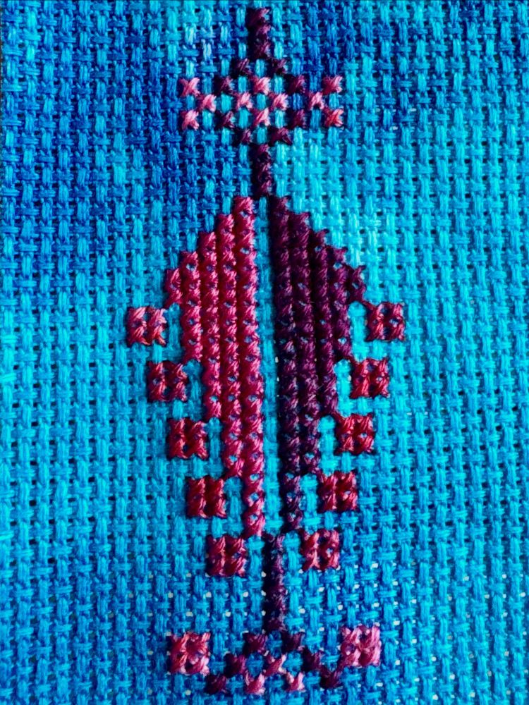 Tatreez, a form of Palestinian folk embroidery, stitched onto blue dyed aida cloth. It shows a red, maroon and pink cypress tree.
