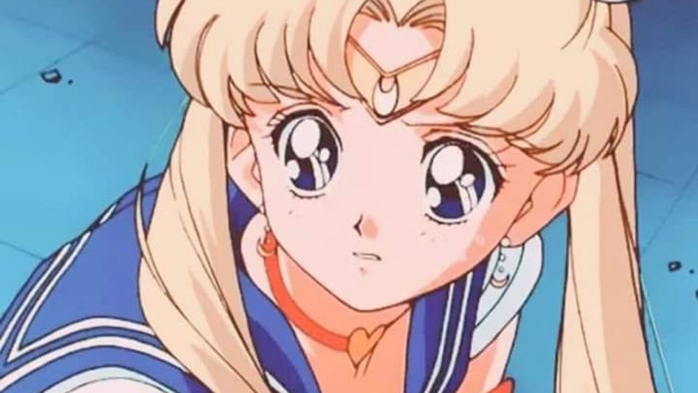 An anime-style illustration of a young girl with blonde hair styled in two buns and flowing pigtails. She has large, expressive blue eyes with a determined yet emotional expression. She wears a blue sailor-style outfit with a red choker featuring a heart pendant, along with a crescent moon headpiece on her forehead. 