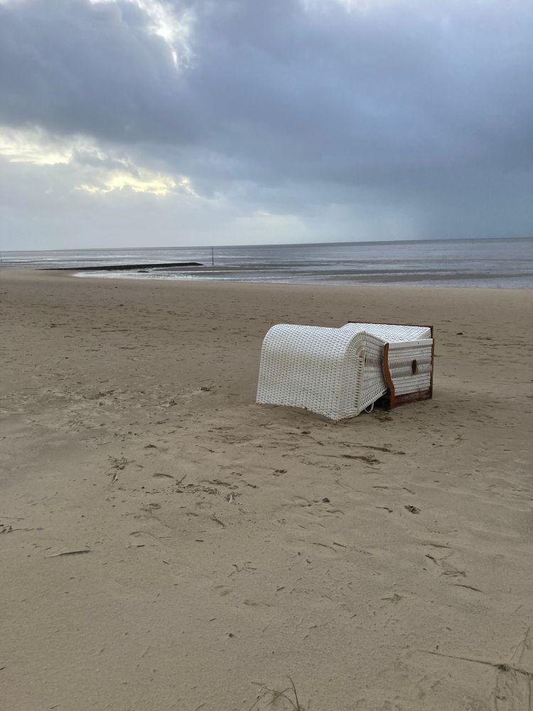Ein vom Sturm umgepusteter Strandkorb liegt mit der offenen Seite nach unten im Sand. Der Strand ist menschenleer, es ist Ebbe. Der Himmel hängt voller schwerer, dunkler Regenwolken. Durch eine Lücke fällt ein bisschen Sonnenlicht.