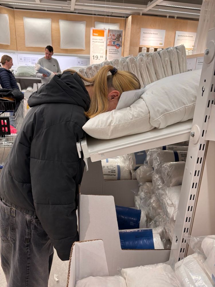 Ein Teenager steht bei IKEA an einem Regal mit Kissen. Sie ist mit dem Gesicht voran in ein Kissen gefallen, das Foto ist von der Seite aufgenommen.