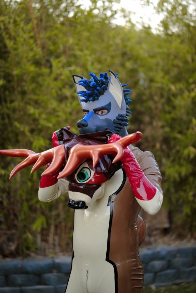 A man in a brown, white and black striped rubber deer suit, taking off an inflatable deer mask, revealing underneath a rubber puppy wolf mask in grey and blue tones 