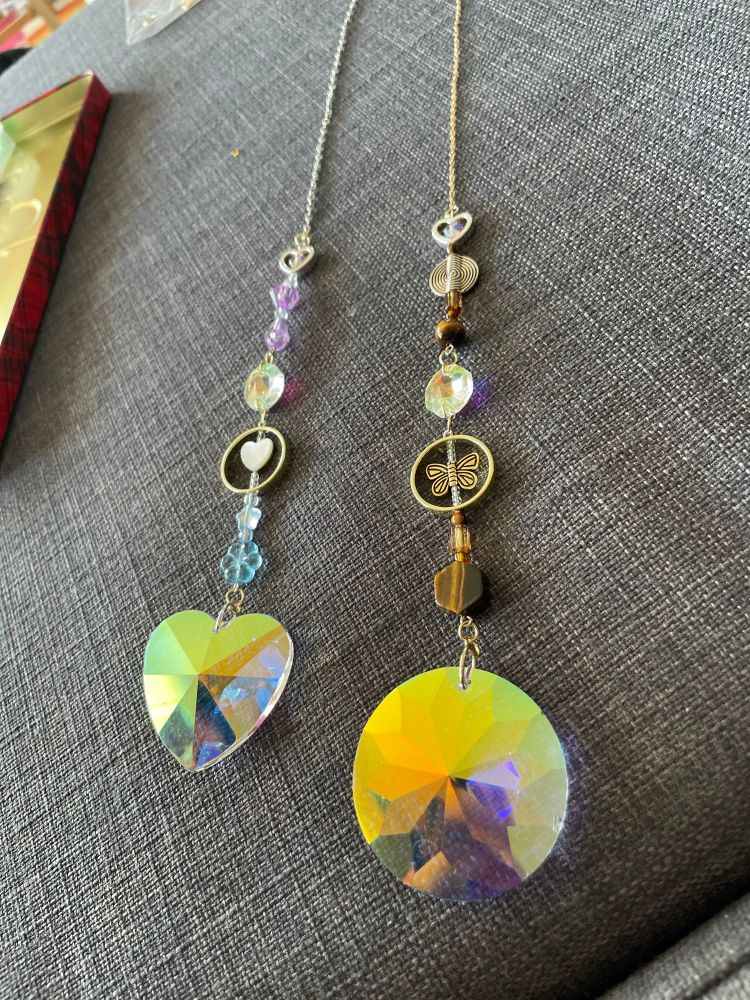 Two suncatchers that I crafted laying side to side on a fabric background. The suncatcher to the left comprising a heart shaped crystal, a smaller heart bead within a circle, as well as various blue and purple beads. The suncatcher to the right comprises a large circle shaped crystal as well as various brown or copper colored beads.